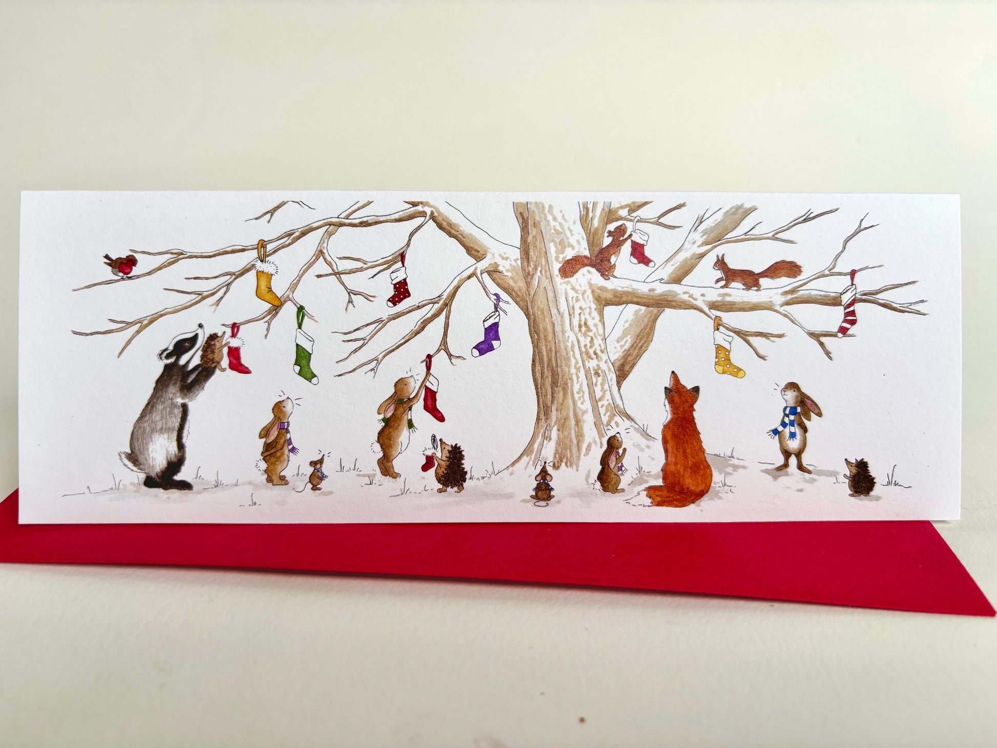 A long slim illustrated christmas card card from my original watercolour showing woodland animals hanging colourful stockings on a bare tree, placed on top of a red envelope against a light neutral background.