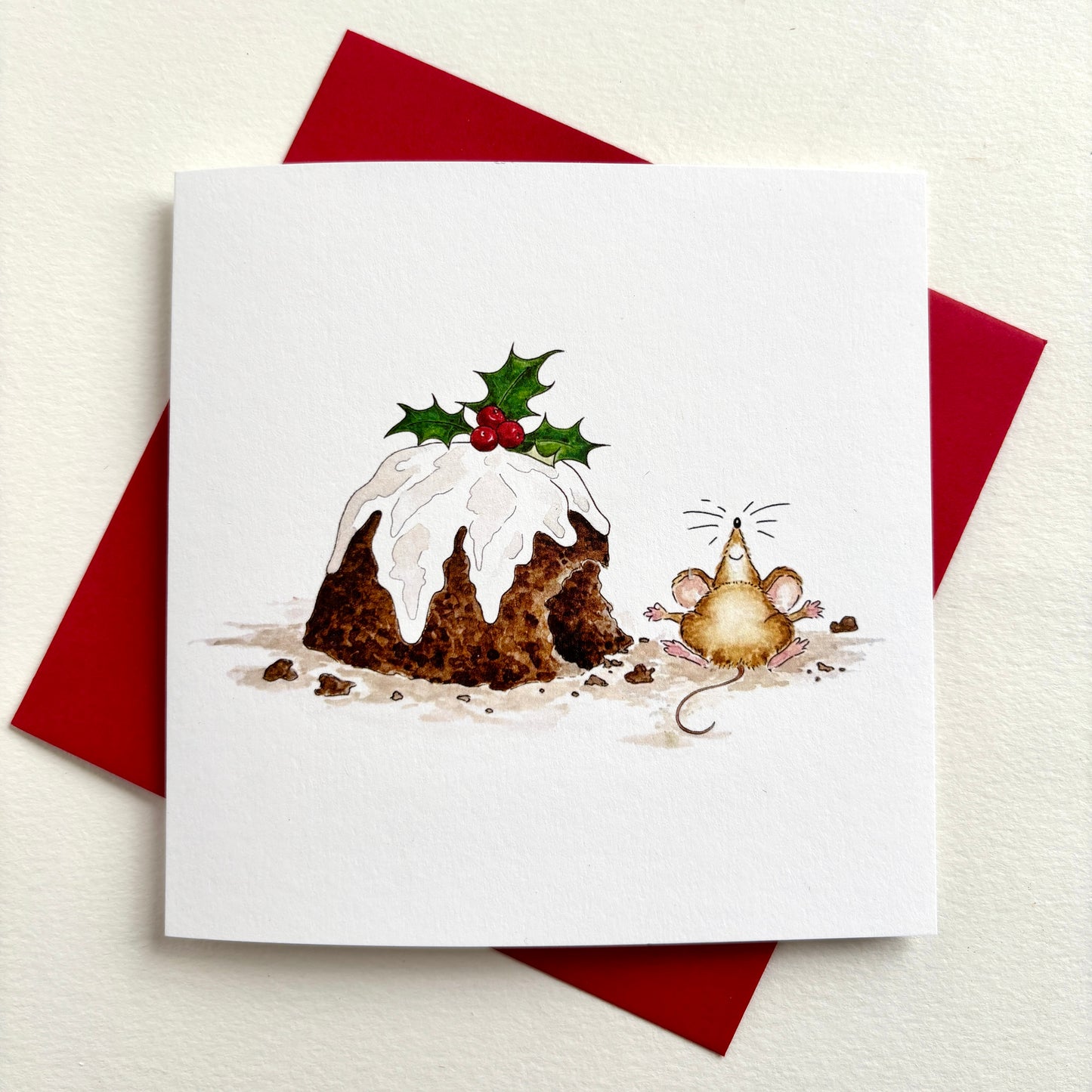 A square charity Christmas card featuring a hand-painted illustration of a Christmas pudding topped with holly and a small mouse beside it. Shown with red envelope.