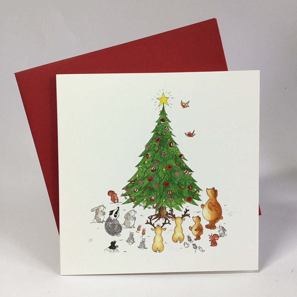 A square white greeting card Christmas card showing a colourful illustration of woodland animals and birds gathered around a decorated Christmas tree topped with a yellow star, shown in front of a red envelope 