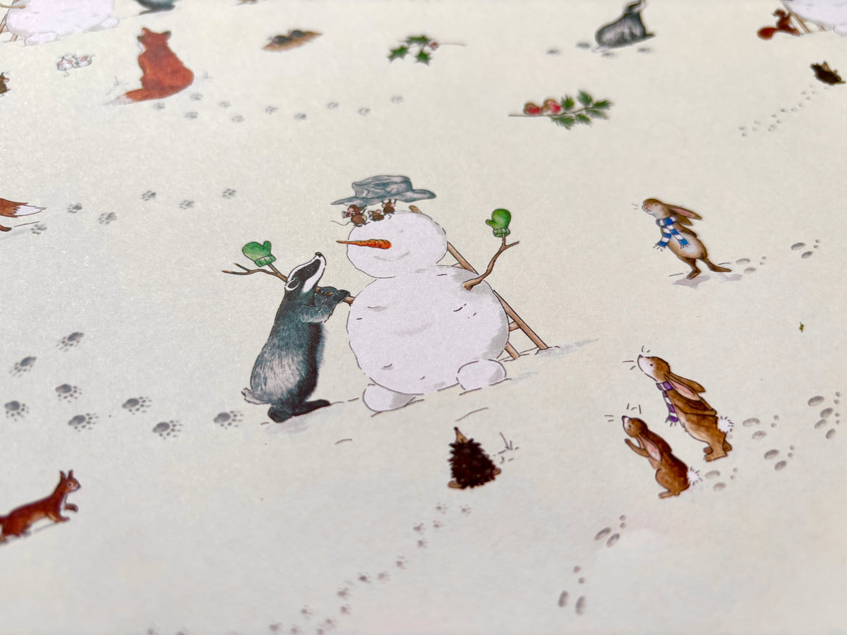 A close-up of sheets of Christmas wrapping paper featuring a winter scene with hedgehogs, badger, rabbits, squirrel and mice building snowman on a warm cream background. From an original al watercolour illustration 